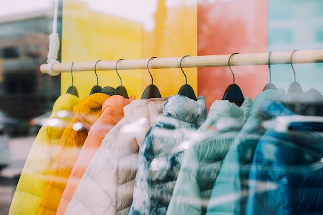 A line of puffer jackets in colors of the rainbow hang on a clothing rack in a display window.
