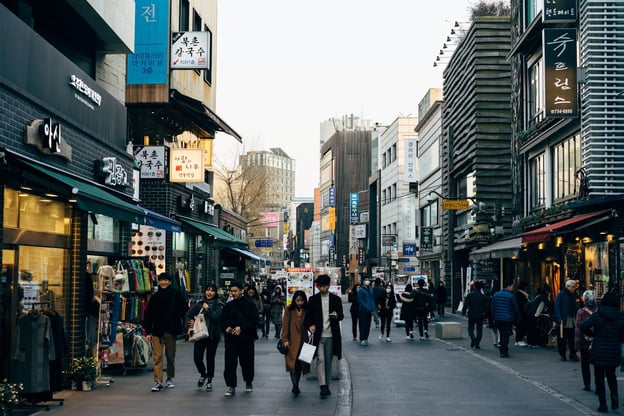 People walk down a crowded street, with shops and signage lining both sides of the road.