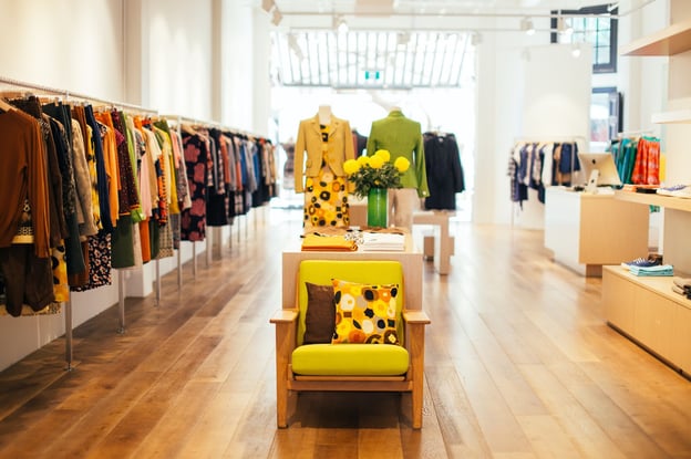 An interior view of a boutique with colorful clothes and an inviting chair in the center of the room.