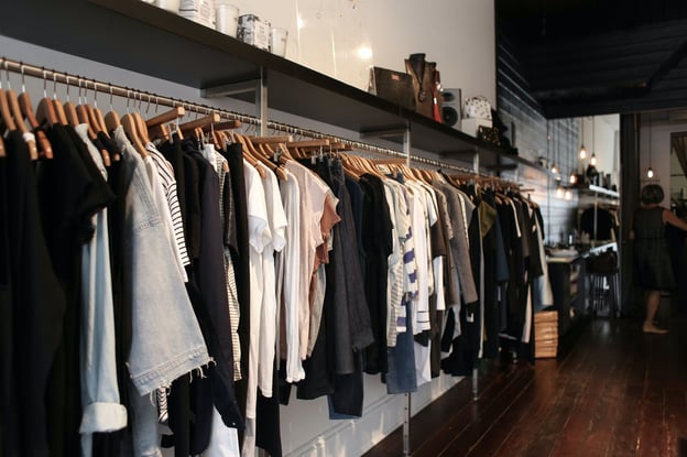 An assortment of women's apparel hangs on a clothing rack lining the back wall of a small boutique fashion shop.