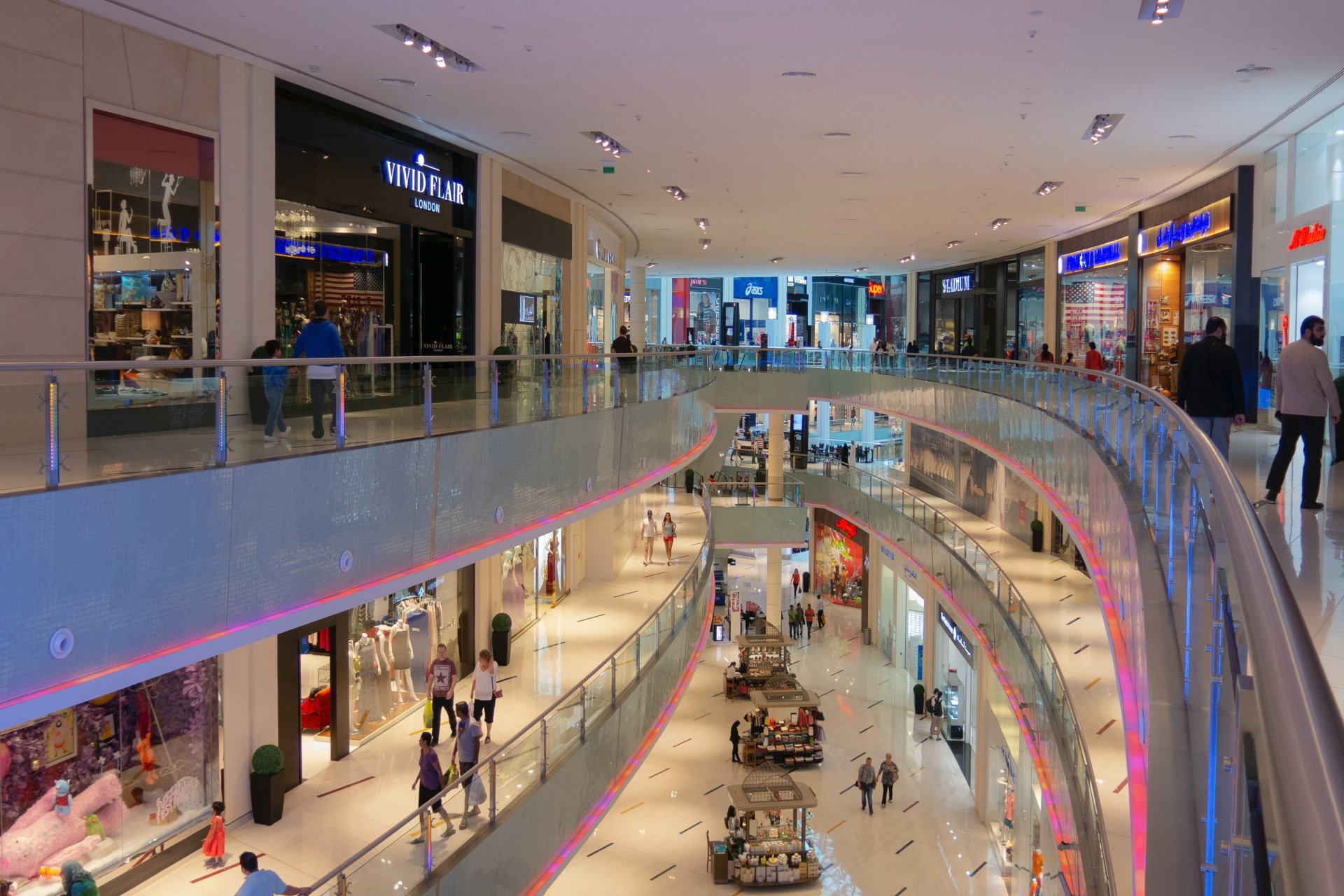 Pedestrians walk through a three-level mall with white ceilings and floors and colorful lighting.