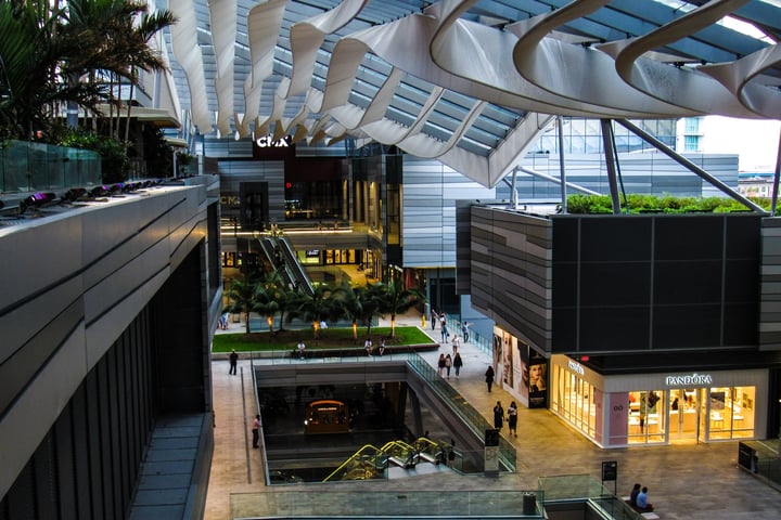 A wide shot of an open-air mall, with glass ceilings letting natural light in to illuminate storefronts, greenery, escalators, and patrons walking about.
